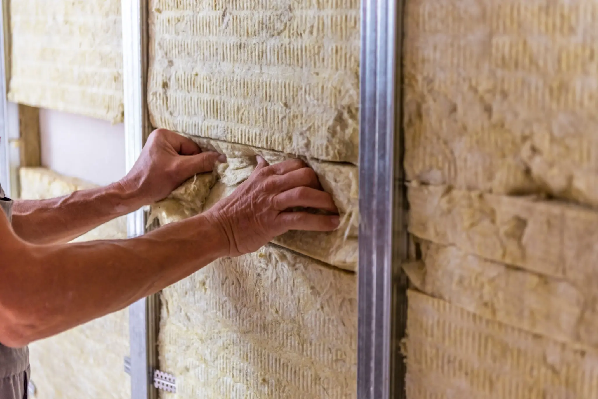 Hands installing yellow insulation material between metal studs in a wall.